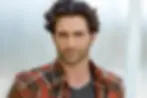 Man with wavy hair and stubble wearing a red plaid shirt and gray tee. Neutral expression, light background.