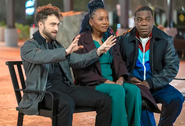 Three people sit on a bench outdoors. The man on the left gestures animatedly. They wear casual jackets, with vibrant colors in the background.