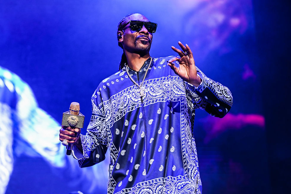 Rapper Snoop Dogg onstage in sunglasses and blue paisley shirt, holding a jeweled microphone under purple concert lights.