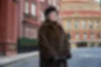 A woman in a fur coat and hat stands on a street with historic buildings in the background. She appears poised, with a neutral expression.