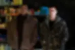 Two serious young men stand in a storeroom with shelves of supplies and boxes behind them, wearing brown and camo jackets.