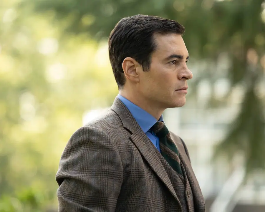 Man in brown plaid suit and blue shirt gazes to the right outside. Blurred green foliage in the background. Serious expression.
