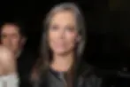 Gray-haired woman in a black jacket smiles at the camera in a dark indoor setting, with two people partially visible behind her.