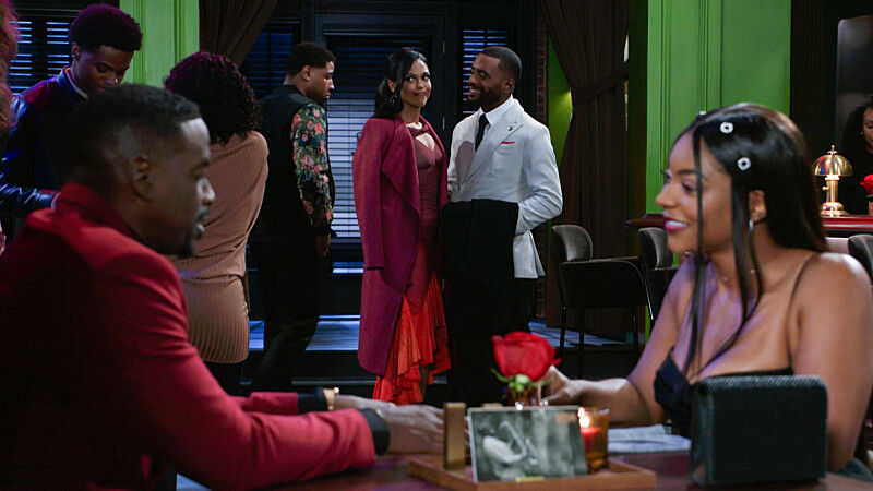 A couple sits at a table holding hands in a dimly lit restaurant. Other patrons socialize in the background. Red and green decor dominates.