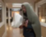 Man in green hooded blanket holding a small dog and a pillow, standing in a well-lit hallway with wooden floors, looking serious.