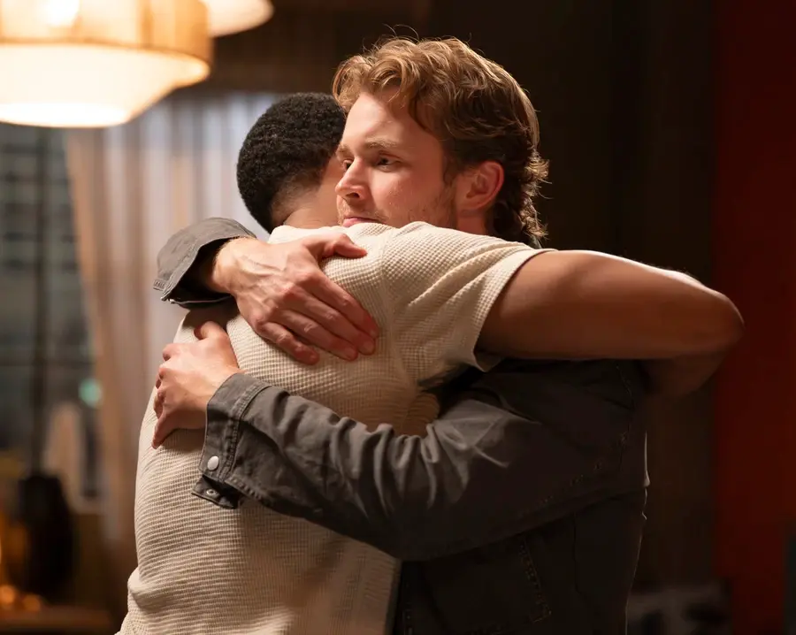 Two people warmly embrace in a softly lit room with curtains and hanging lamps. Both wear jackets, conveying a comforting and emotional mood.