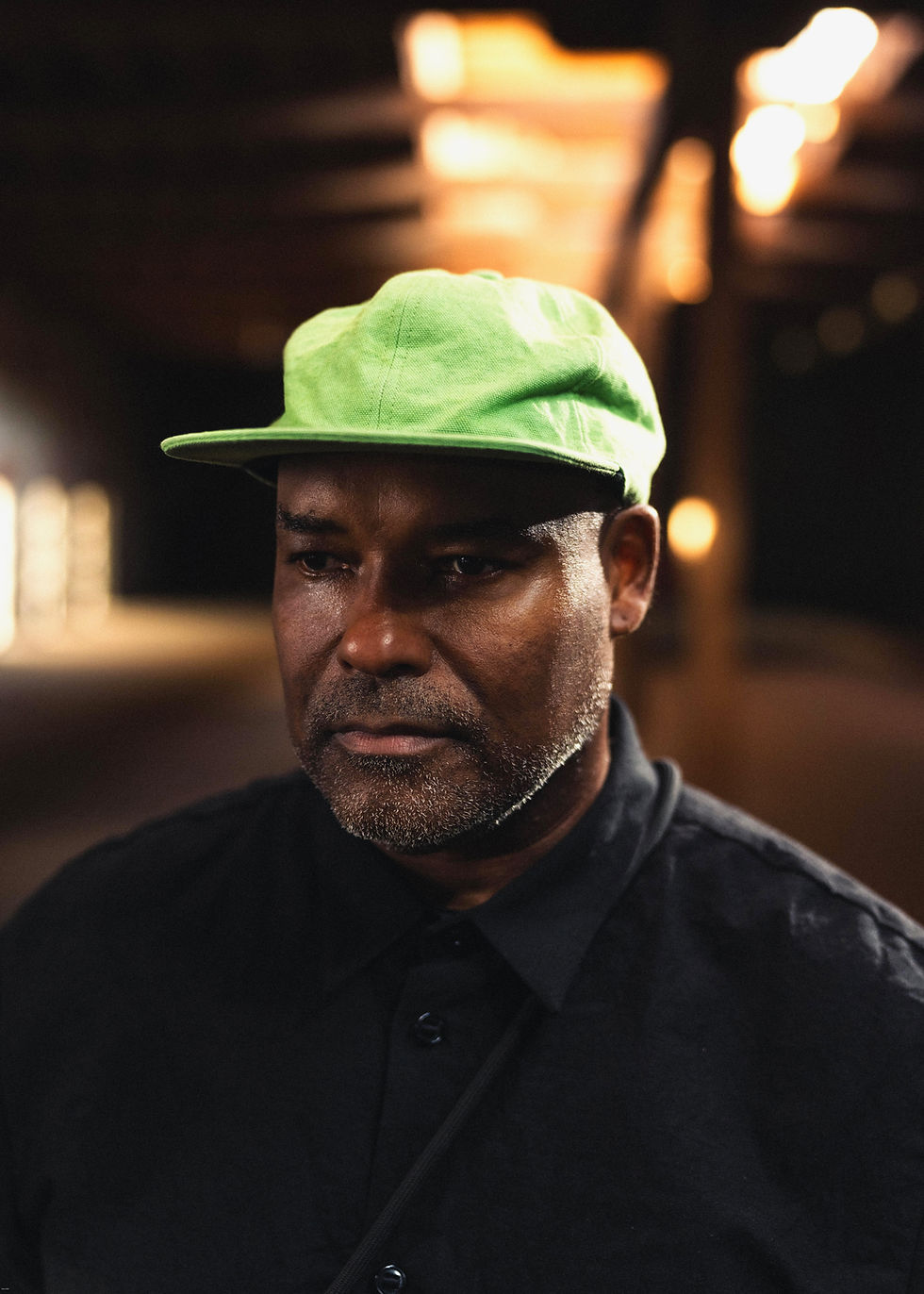 Portrait of a serious man in a bright green cap and dark shirt, lit by warm blurred lights in the background.