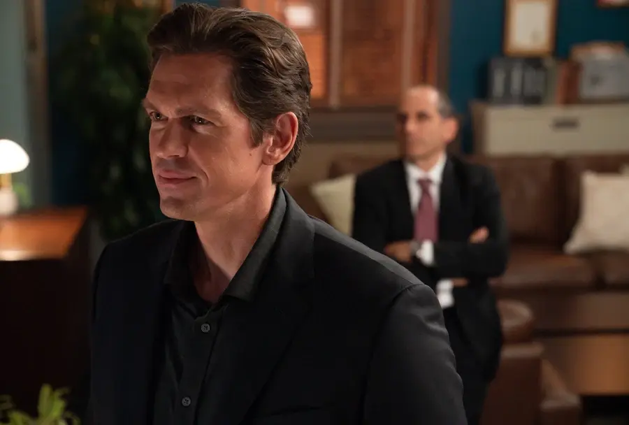 Man in a dark suit smiles confidently in the foreground. Another man sits in the background with arms crossed in an office setting.