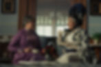 Two women in elegant attire sit on a couch in a vintage room, drinking tea. One wears a purple dress, the other a floral outfit. Warm ambiance.