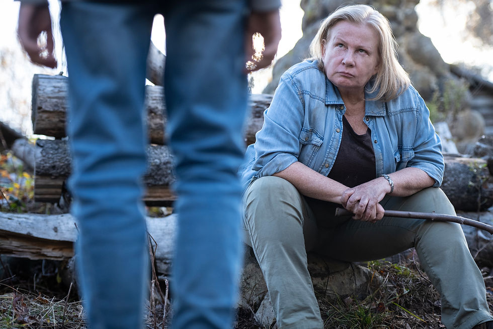 Blonde woman in denim sits on logs outdoors, holding a stick and looking up at a standing person in jeans with a serious expression