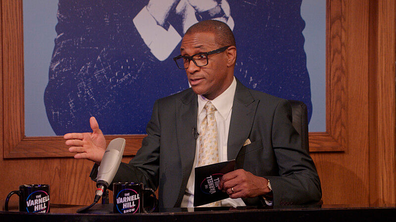 Man in suit and glasses gestures at a talk-show desk, holding a card beside mugs labeled The Varnell Hill Show.