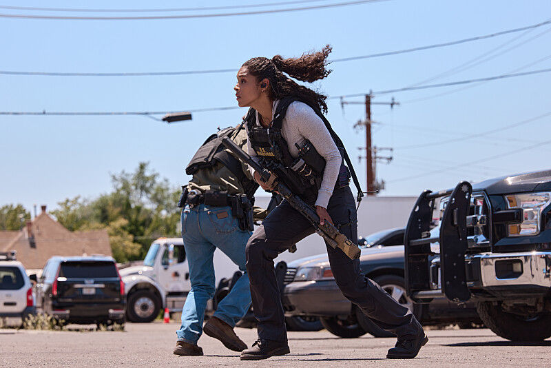 Two tactical officers with rifles run through a sunny parking lot beside pickup trucks and police vehicles.