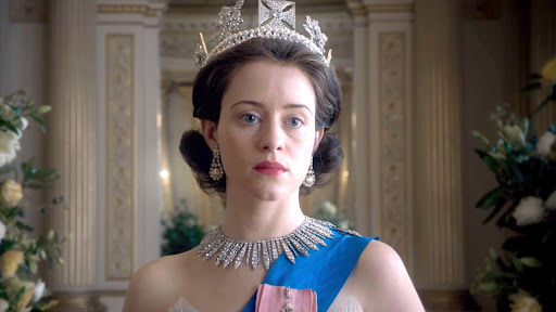 Woman in regal attire stands in ornate room, wearing a crown and necklace. Her expression is serious. Flowers decorate the background.