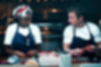 Two cooks in white shirts and navy aprons prep lobster in a dim restaurant kitchen, exchanging focused looks.