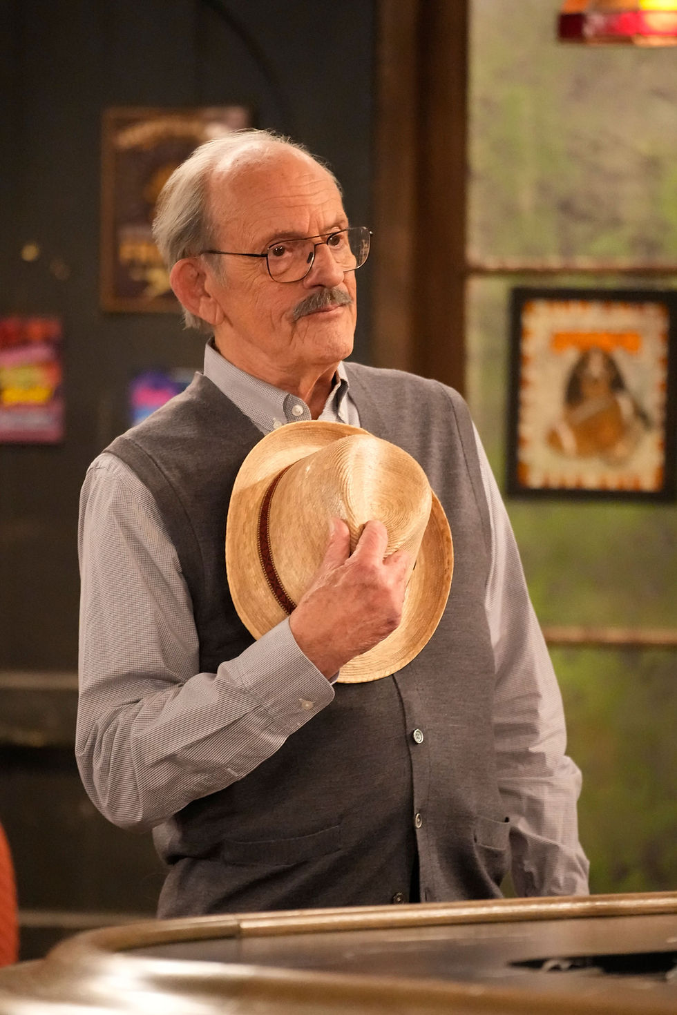 Elderly man with glasses holding a straw hat indoors, wearing a gray vest. Wooden bar and colorful posters in the background.