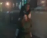 Two women in a dim warehouse; one restrains the other, who appears distressed. Industrial background with labeled pallets; tense atmosphere.