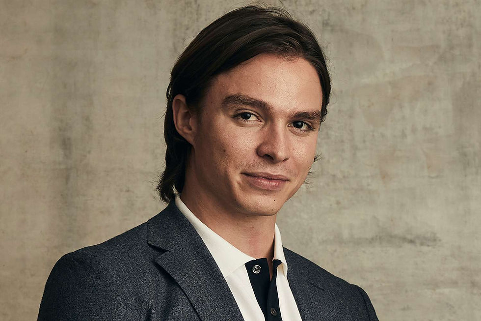 Portrait of a young man in a dark suit and white shirt, слегка smiling against a beige textured wall.