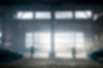 Two figures stand in an old warehouse, pointing guns at each other. Sunlight streams through large windows, illuminating the dusty space.