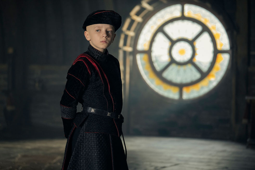 Boy in medieval attire with black and red details stands indoors by a large circular stained glass window, looking thoughtful.