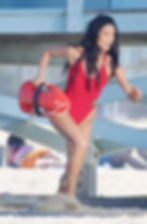 Woman in red swimsuit runs on beach, holding red rescue float. Beachgoers in background. Determined expression.