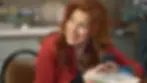 Smiling woman with long red hair holds a phone in a cozy kitchen setting. She's in a red jacket, creating a warm, cheerful mood.