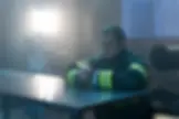 Firefighter in uniform sits at a metal table with hands clasped, in a dim, smoky room. A bright light glows behind, creating a somber mood.