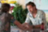 Two men in a bar converse, one with a floral shirt leans on the counter. A "Wash Your Hands" sign and beer bottle are visible. Plant background.