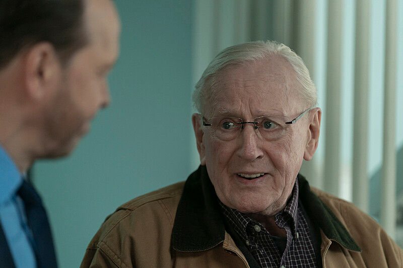 Elderly man in glasses wearing a brown jacket speaks to another person. They're indoors with light blue walls and vertical blinds.