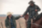 Two cowboys in hats exchange a handshake across a barbed-wire fence in a muted field, one mounted on a brown horse.