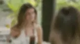 Smiling woman in a white top talks at an outdoor café, with iced drinks on the table and greenery behind her.