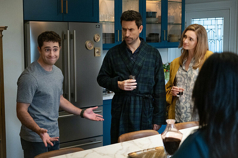Three adults stand in a blue kitchen, two holding drinks while a man gestures awkwardly by the fridge, with a tense mood.