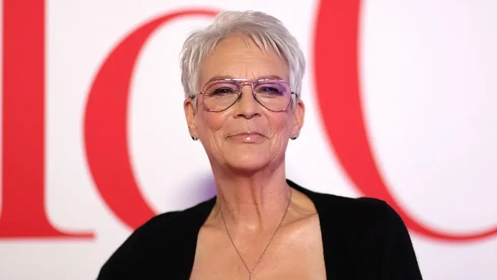 Smiling person with short gray hair and glasses, wearing a black top against a backdrop with large red letters.