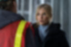 A woman with blonde hair in a dark coat looks serious while talking to a person in a construction vest inside an industrial setting.