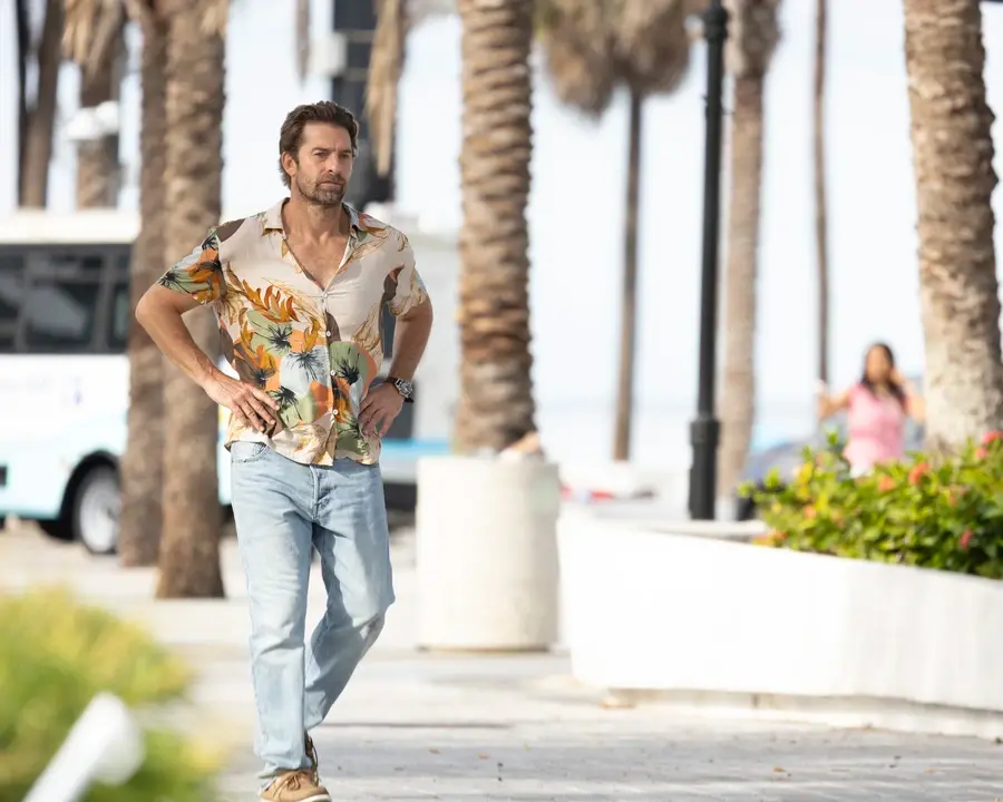 Man in tropical shirt and jeans walks on palm-lined street, hands on hips. Background features trees, bus, and a woman. Sunny mood.