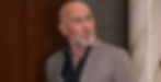 Bald gray-bearded man in a tan blazer looks off to the side indoors, with a thoughtful, serious expression.