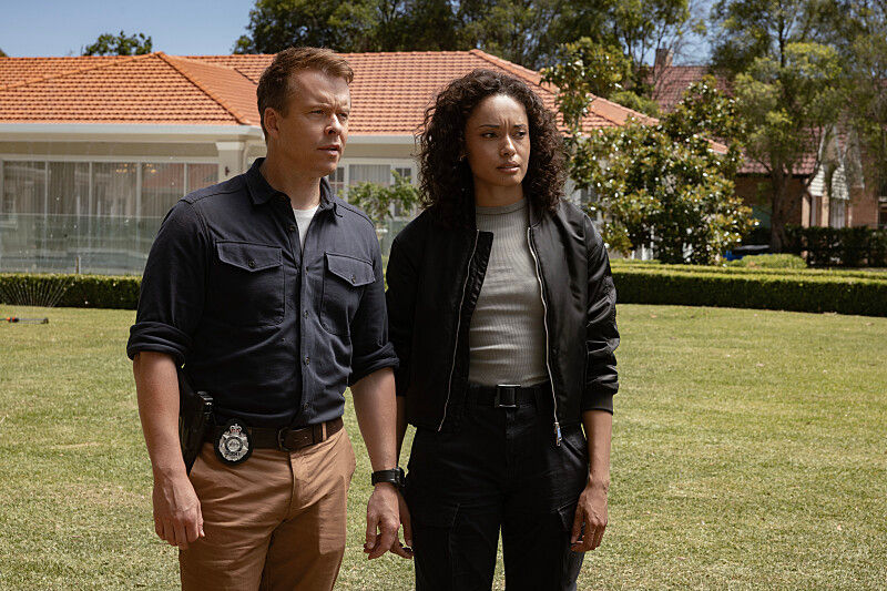 Two detectives stand on a lawn in front of a suburban house, looking serious under a red-tile roof.