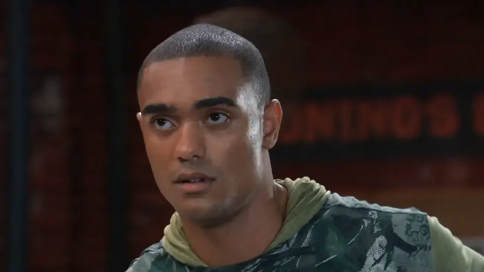Close-up of a serious young man in a green hoodie and camo shirt, against a dark blurred background.