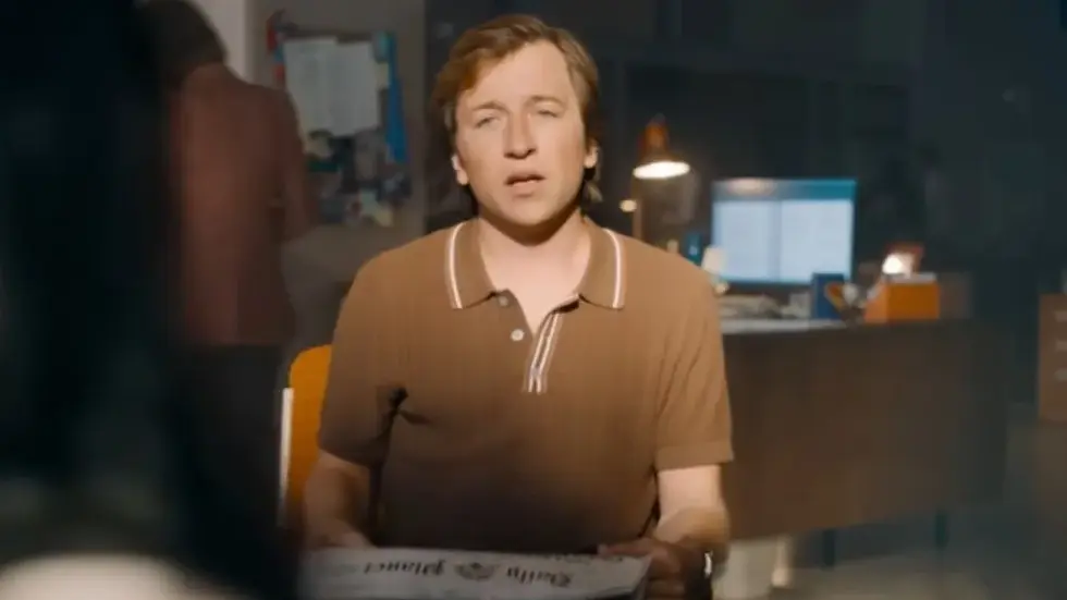Man in brown shirt holds newspaper in office setting, looking towards the camera. Background shows desk with lit computer screens.