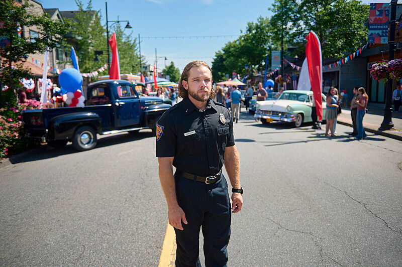 Police officer stands in a sunny parade street with vintage trucks, balloons, and flags; Edgewater banner in background.