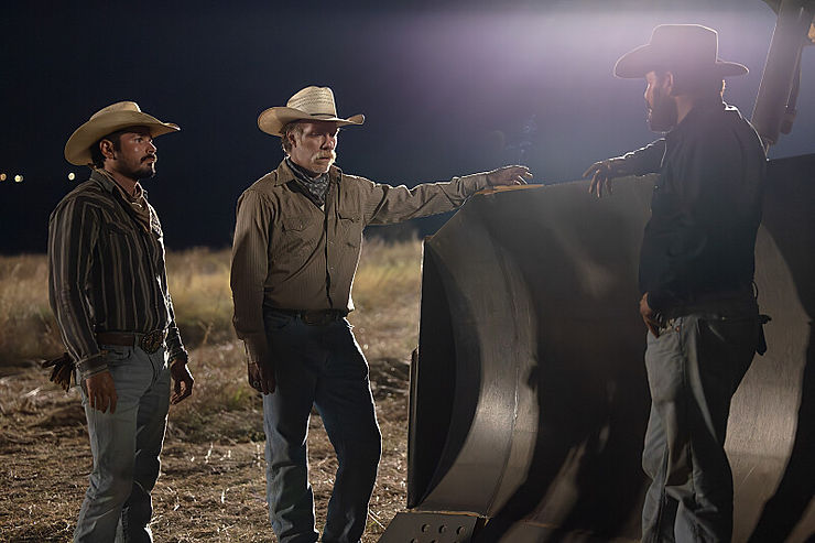 Three cowboys stand in a dim field at night, one pointing at a large metal structure while the others listen under bright lights.