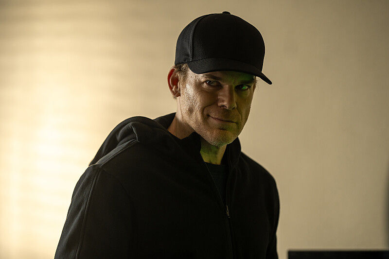 Man in a black cap and hoodie smiles subtly in a dimly lit room with a warm yellow background, creating a mysterious mood.
