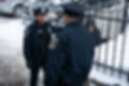 Two police officers converse in a snowy setting while a person behind a snow-covered fence observes. Badge reads "Boston Police."