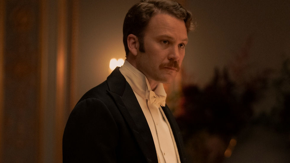 Man in formal attire with a serious expression in a dimly lit room, warm lighting, soft-focus background with floral decor.