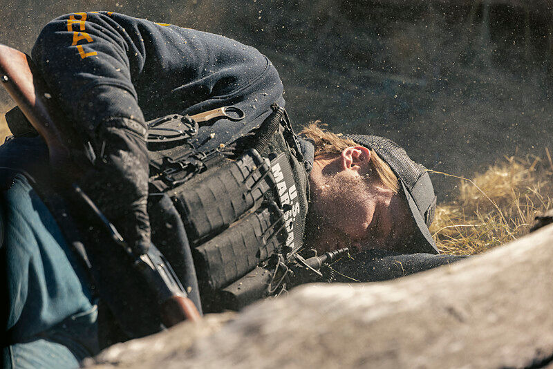 Tactical man in black vest and cap lies prone outdoors in dry grass, aiming a rifle; dust hangs in the air.