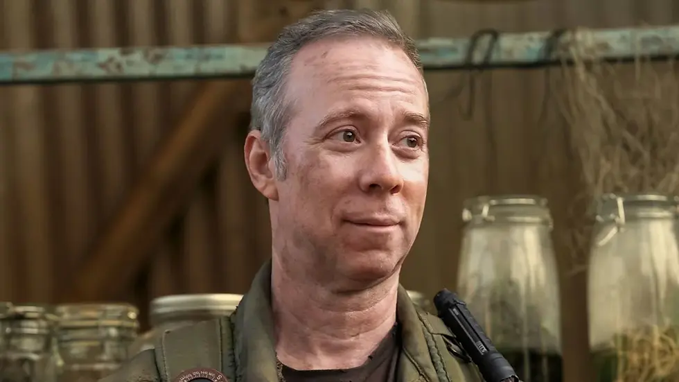 Middle-aged man in military gear smiles in a rustic shed with glass jars and wooden beams behind him.