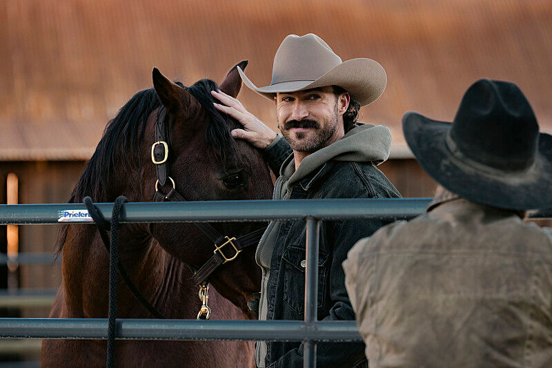 Cowboy in a tan hat pets a brown horse over a metal fence. Another figure in a dark hat watches. Rustic barn setting. Relaxed mood.