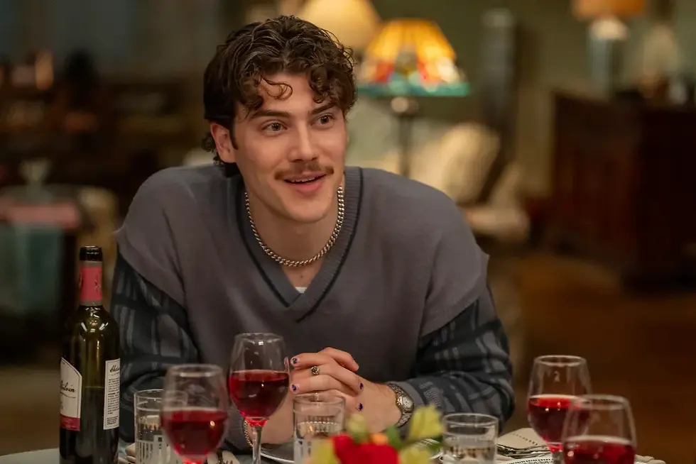 Smiling young man with mustache sits at a dinner table with wine glasses and a bottle in a warm, softly lit room.