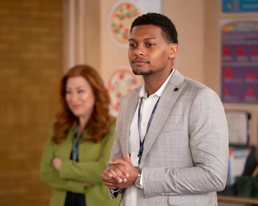 Man in a light gray suit stands confidently, hands clasped. Woman in green blurred in background. Classroom posters on walls.