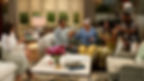 Five people sitting on couches in a cozy living room, raising glasses and smiling. Bright flowers and books are on a central table.