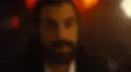 Bearded man with long hair in a dim, warmly lit room. Intense expression, with red and orange lights in the blurred background.